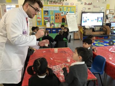 Ryan (dark hair with dark glasses and wearing a white lab coat) is standing talking to three uniformed children seated at a table. The table has a red table cloth with slime making materials on it.